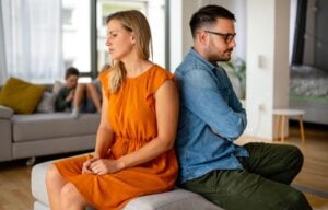 Unhappy couple sitting back-to-back in living room while child plays in background, representing divorce challenges.