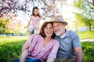 Grandparents smiling outdoors with their granddaughter playing in the background.